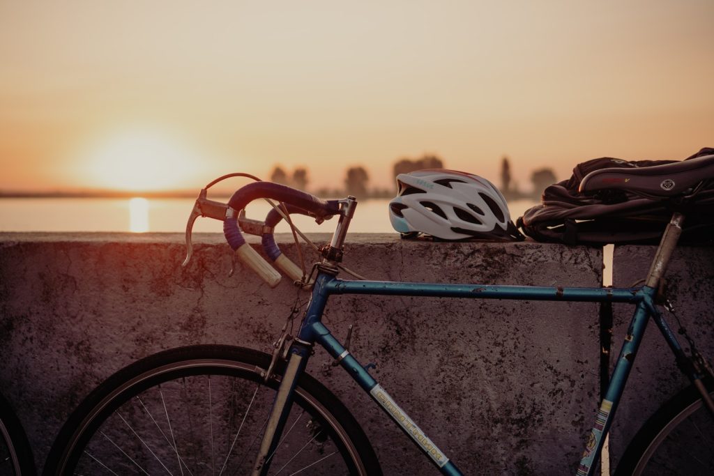 Bicicleta con manubrio deportivo contra una pared donde se apoya un casco y una mochila y de fondo el atardecer al horizonte