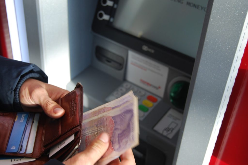 Mano sosteniendo billetera marrón con billetes frente a un cajero Mano sosteniendo billetera marrón con billetes frente a un cajero