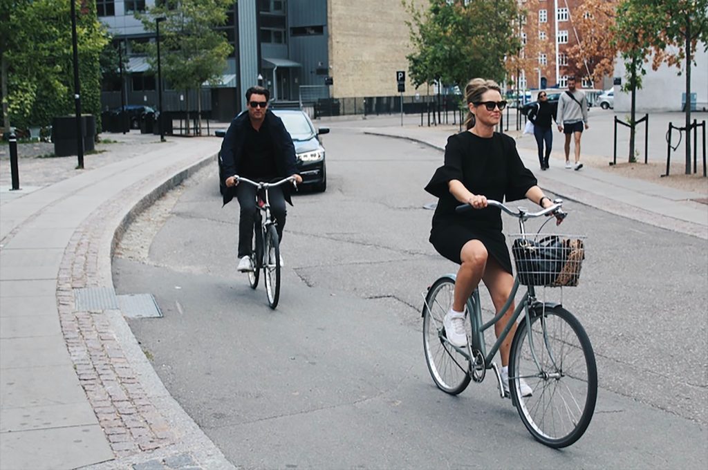 Mujer en vestido negro andando en bicicleta seguida de hombre también en bicicleta, andando sobre una calle
